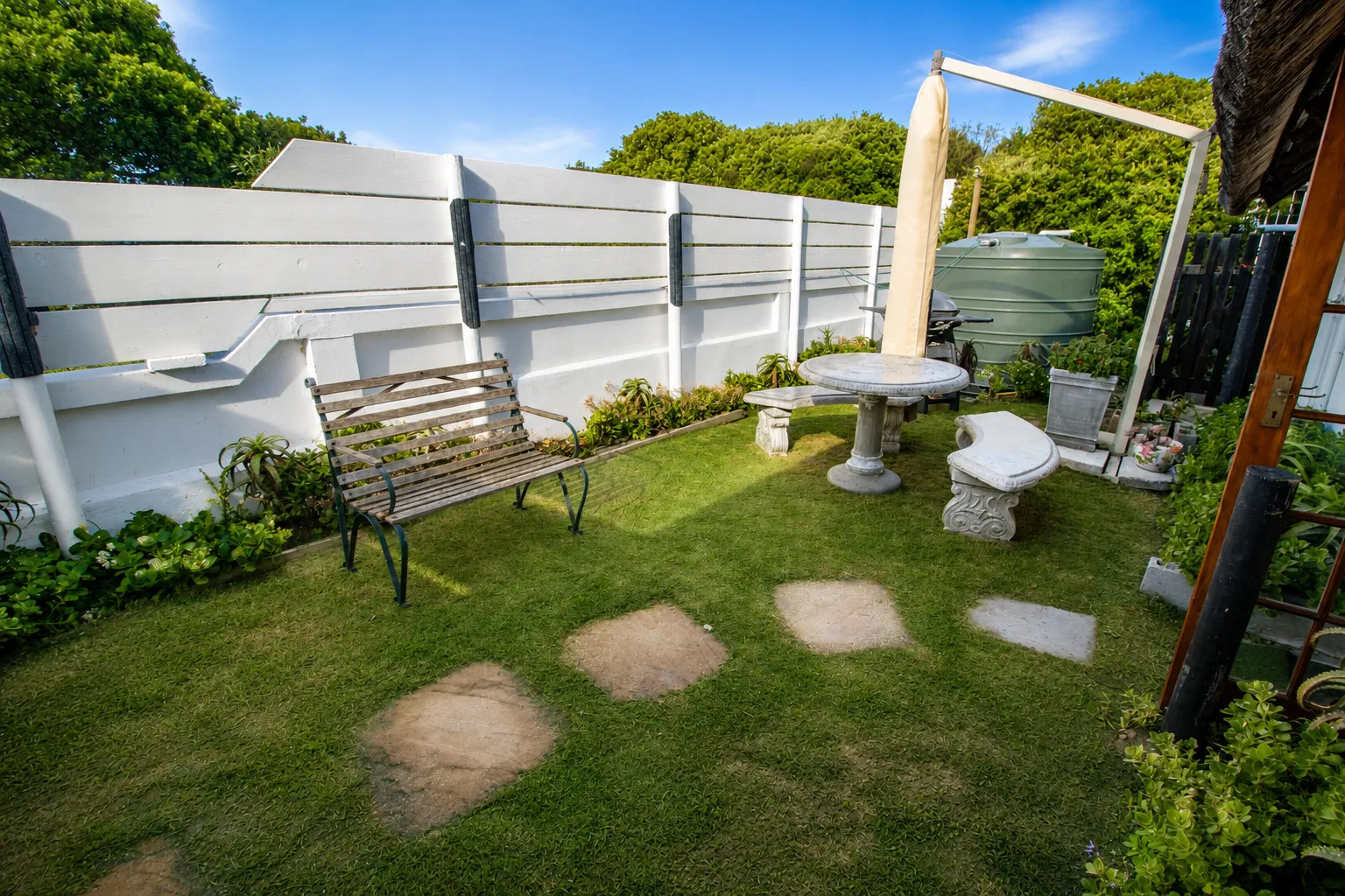 The side garden lawn with a stone table and benches, backed by a rainwater tank and white slatted fence